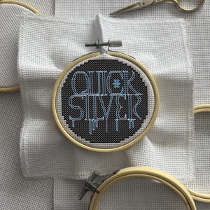 May include: A cross-stitch embroidery hoop with the words "QUICKSILVER" in a blue and white font. The design is on a black background with a snowflake symbol. The hoop is set on white fabric, with other hoops and scissors in the background.