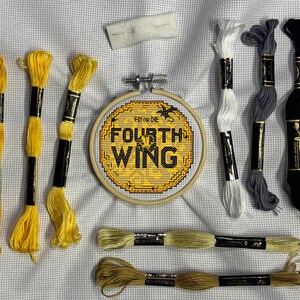 May include: A cross-stitch project with the text "FOURTH WING" in yellow and black, set in a wooden hoop. Several skeins of embroidery floss in yellow, white, gray, and black are arranged around the hoop, along with a needle and felt. The text "FLY OR DIE" is above the main text.