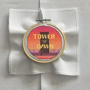 May include: A cross-stitch embroidery hoop featuring the words "TOWER OF DAWN" in gold lettering. The design includes a black tower and a sunset-colored background. The embroidery is set against a white fabric.