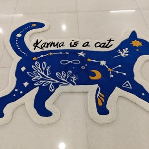 以下が含まれることがあります： 白い縁取りのある青い猫の形をした敷物。敷物は白い背景に青い猫のシルエットがあり、星、三日月、無限大記号など、さまざまな天体模様が描かれています。敷物には「Karma is a cat」という文字が黒で書かれています。