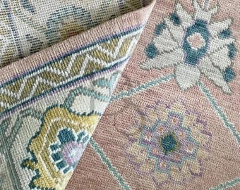 May include: A pink and cream rug with a floral pattern. The rug has a detailed border with a geometric design. The rug is folded in half, showing the back side.