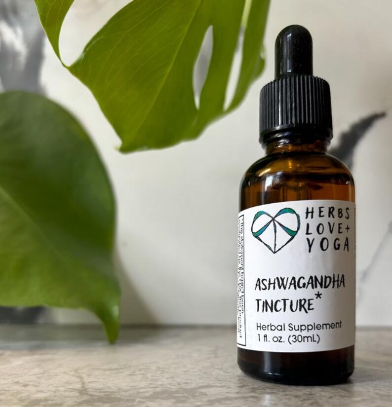 May include: A brown glass bottle with a black dropper and a white label. The label reads "HERBS LOVE+ YOGA" and "ASHWAGANDHA TINCTURE* Herbal Supplement 1 fl. oz. (30ml)". A green leaf is in the background.