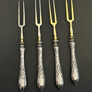 May include: Four antique silver forks with ornate handles and two-pronged tines. Two forks have silver tines, and two have gold-colored tines. The handles feature a floral design.