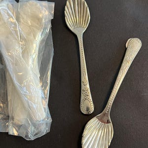 May include: Silver-colored spoons with a shell-shaped bowl and ornate handles. One spoon is out of its plastic packaging, and another is displayed separately. The spoons are likely for serving or eating.