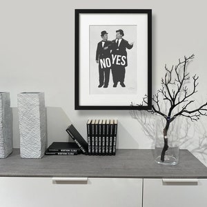 Puede incluir: Fotograf&iacute;a en blanco y negro enmarcada de dos hombres sosteniendo un cartel que dice "NO YES". La foto est&aacute; sobre una encimera gris con dos jarrones grises texturizados, un jarr&oacute;n con una rama negra y una pila de libros negros.