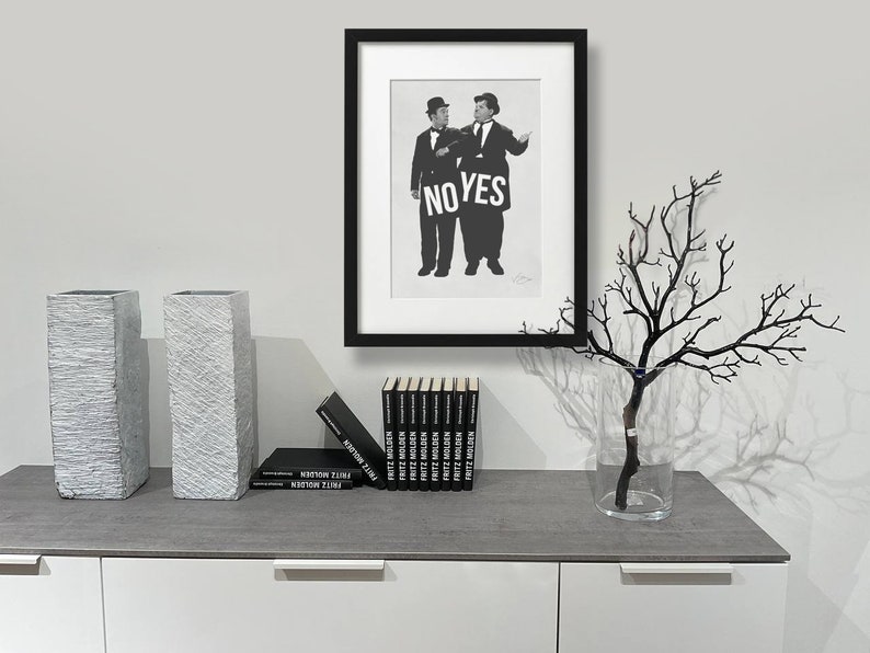 Puede incluir: Fotograf&iacute;a en blanco y negro enmarcada de dos hombres sosteniendo un cartel que dice "NO YES". La foto est&aacute; sobre una encimera gris con dos jarrones grises texturizados, un jarr&oacute;n con una rama negra y una pila de libros negros.