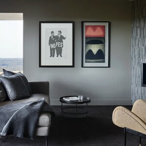 Puede incluir: Una sala de estar con un sof&aacute; gris, una mesa de caf&eacute; negra y un sill&oacute;n beige. Dos cuadros enmarcados cuelgan de la pared. Una imagen muestra a dos hombres sosteniendo un cartel que dice "NO YES". La otra imagen es un dise&ntilde;o abstracto con colores rojo, negro y blanco.