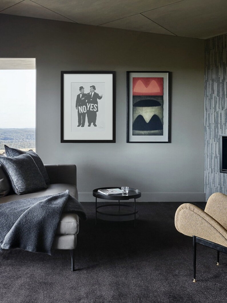 Puede incluir: Una sala de estar con un sof&aacute; gris, una mesa de caf&eacute; negra y un sill&oacute;n beige. Dos cuadros enmarcados cuelgan de la pared. Una imagen muestra a dos hombres sosteniendo un cartel que dice "NO YES". La otra imagen es un dise&ntilde;o abstracto con colores rojo, negro y blanco.