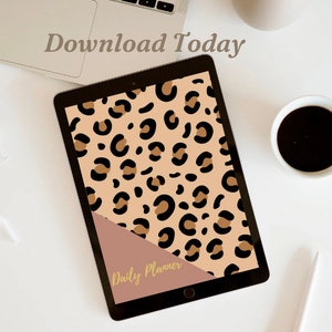 May include: A digital tablet with a black frame and a screen displaying a leopard print pattern. The screen is divided diagonally with a brown section at the bottom that says "Daily Planner" in gold lettering. The tablet is on a white surface with a laptop, a cup of coffee, and a pen.