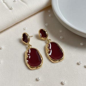 Peut inclure: Une paire de boucles d'oreilles pendantes dorées avec un remplissage en émail rouge foncé. Les boucles d'oreilles ont une forme irrégulière et organique avec une bordure dorée. Elles sont présentées sur une surface blanche avec de petites perles blanches.