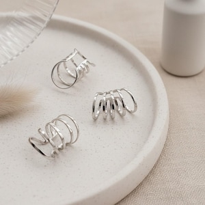 Pode incluir: Três brincos de orelha prateados são exibidos em uma bandeja branca salpicada. Os brincos de orelha têm um design minimalista com múltiplas faixas curvas. O fundo inclui uma garrafa branca e uma superfície macia de tom neutro.