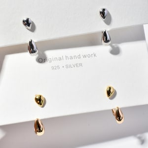 Op de afbeelding: Een verzameling zilver- en goudkleurige druppelvormige oorbellen. De oorbellen worden gepresenteerd op een witte kaart met de tekst "Original hand work 925 SILVER". De oorbellen hebben een gladde, gepolijste afwerking en een eenvoudig, elegant ontwerp.