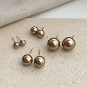 Può includere: Diverse paia di orecchini a perno con perle dorate di varie dimensioni sono disposti su una superficie neutra. Gli orecchini presentano un classico design a perla rotonda con perni dorati. L'illuminazione mette in risalto la lucentezza delle perle.