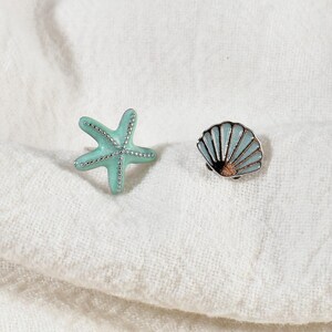 May include: A pair of ocean-themed earrings. One earring is a light blue starfish with silver accents. The other is a light blue seashell with a silver border and a brown center. Both earrings are on a white, textured surface.