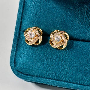 May include: A pair of gold-colored stud earrings. Each earring features a central clear gemstone surrounded by smaller clear gemstones and a woven gold-tone design. The earrings are displayed on a teal velvet surface.