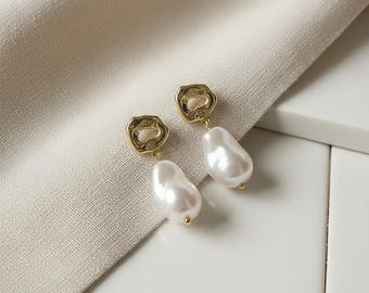Könnte beinhalten: Ein Paar goldfarbene Ohrringe mit einem einzigartigen Design. Jeder Ohrring hat eine strukturierte, unregelmäßige goldene Oberseite mit einem hängenden, großen, weißen, perlmuttartigen Tropfen. Die Ohrringe werden auf einem cremefarbenen Stoff präsentiert.