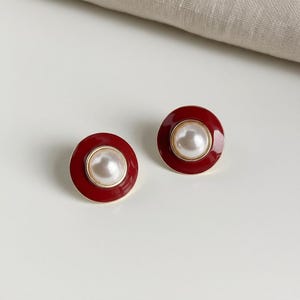May include: A pair of round stud earrings. Each earring features a large, off-white pearl center, surrounded by a gold-colored ring, and a deep red enamel border. The earrings are set against a white background.