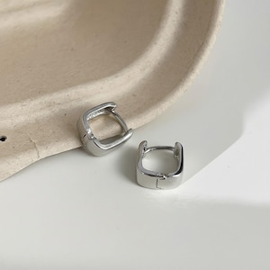 Peut inclure: Une paire de petites créoles argentées de forme carrée. Ces boucles d'oreilles présentent une finition métallique polie et réfléchissante ainsi qu'un mécanisme de fermeture à charnière, conçues pour se porter près du lobe de l'oreille.