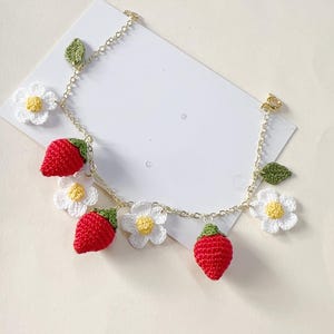 Peut inclure: Un bracelet en chaîne dorée avec des breloques en crochet en forme de fraises et de marguerites. Les breloques sont rouges, blanches et vertes.