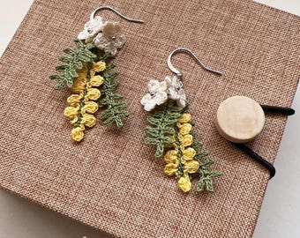 May include: Crocheted earrings featuring yellow and green mimosa flowers with white flower accents. The earrings are hanging from silver hooks.