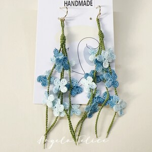 Pode incluir: Um par de brincos feitos à mão de crochê com flores azuis e verdes. Os brincos são feitos com uma técnica de crochê delicada e são perfeitos para adicionar um toque de fantasia a qualquer roupa. Os brincos são etiquetados como "Uncinetto Handmade".