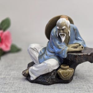 Peut inclure: Une figurine en céramique représentant une personne âgée à barbe et cheveux blancs, vêtue d'une robe bleue et d'un pantalon blanc. Le personnage porte un chapeau à larges bords, s'appuie sur une base rocheuse brune et un petit pot en céramique.