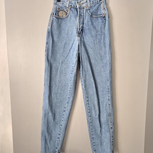 May include: High-waisted light blue denim jeans featuring a button fly closure, five pockets, and a small rectangular patch with the text "MANAGE" on the right front pocket. The jeans have a tapered leg silhouette and copper-coloured metal rivets.
