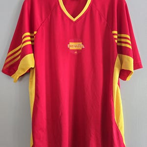 May include: Red and yellow soccer jersey with a V-neck collar. The jersey features the word "ESPAÑA" and the Adidas logo. Yellow accents are on the sleeves and sides. The jersey is hanging on a hanger.