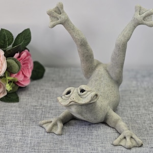 May include: A light grey, speckled frog figurine in a handstand position with its front legs extended forward and back legs pointing upwards. The frog has a wide, flat head with dark eyes and webbed feet, crafted with a textured, stone-like finish.