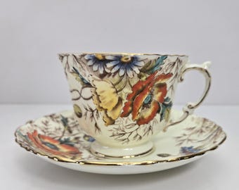 Può includere: Tazza da tè e piattino color crema con motivo floreale. La tazza e il piattino presentano fiori rossi, gialli e blu. Bordi dorati. Un classico set da tè per servire il tè.