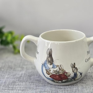 May include: A white ceramic two-handled mug featuring a colorful illustration of Peter Rabbit and other rabbits. The text on the mug includes the words four, Flopsy, and Cotton-tail. The mug has a rounded body and two symmetrical side handles.
