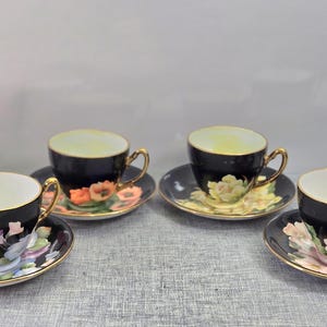 Peut inclure: Quatre tasses à thé en porcelaine noire avec soucoupes assorties, chacune ornée d'un motif floral unique dans des tons de rose, orange, jaune et violet. Les tasses et soucoupes ont des bords et des anses dorés, avec un intérieur blanc.