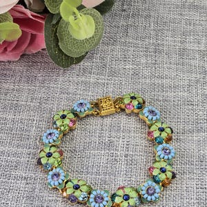 May include: A gold bracelet with a floral design. Each flower has a light blue enamel base, a central purple gem, and colourful gems. The bracelet is connected with a decorative clasp. A beautiful piece of jewellery.