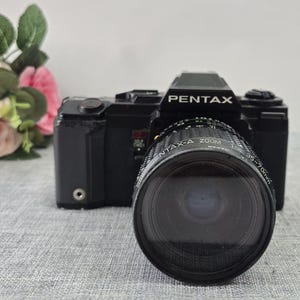 以下が含まれることがあります： Pentax-A Zoom 35-70mmレンズを装着した黒いPentax一眼レフカメラ。カメラ本体のプリズムハウジング上部にはPentaxのロゴがあり、前面にはセルフタイマーのスイッチが配置されています。レンズ鏡筒には白い文字で技術仕様が記載されています。