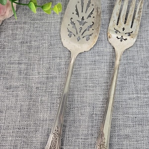 May include: A vintage silver serving spoon and fork set. The spoon has a decorative pierced design, and the fork has a similar etched pattern. Both utensils have detailed floral accents on the handles. Perfect for serving at a formal dinner.