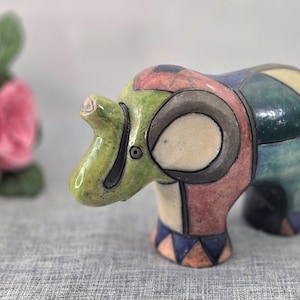 May include: A ceramic elephant figurine featuring a segmented, multi-colored geometric pattern in shades of green, pink, blue, and cream. The surface is divided by dark outlines, creating a mosaic-like appearance across the body and legs.
