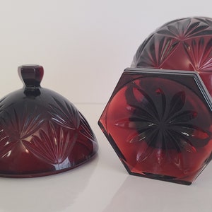 May include: A red glass candy dish with a lid. The dish has a hexagonal base and a round top with a geometric pattern. The lid has a knob on top.