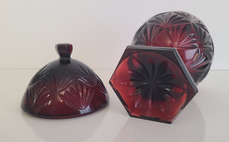 May include: A red glass candy dish with a lid. The dish has a hexagonal base and a round top with a geometric pattern. The lid has a knob on top.