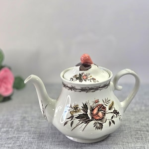 Op de afbeelding: Een witte keramische theepot met een bloemmotief van roze rozen en donkerbruin blad. Het deksel is bekroond met een roze knop en versierd met kleine roosjes. Een donkerbruine decoratieve rand loopt rondom de bovenkant van de theepot.