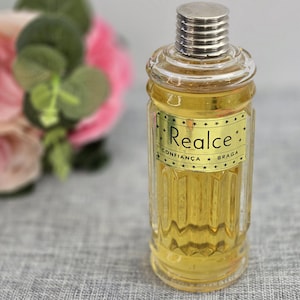 May include: A clear glass perfume bottle with a silver cap and a gold label that reads "Realce Confianca Braga". The bottle is filled with a yellow liquid. A blurred bouquet of pink and green flowers is in the background.