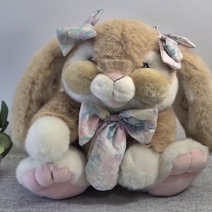 May include: A plush stuffed rabbit with soft tan and white fur, featuring long floppy ears adorned with two pastel floral bows. The rabbit has a large matching floral bow around its neck, a pink nose, and light pink paw pads on its feet.