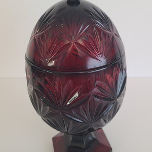 May include: A red glass candy dish with a lid. The dish is shaped like an egg and has a geometric pattern. The dish is on a pedestal.