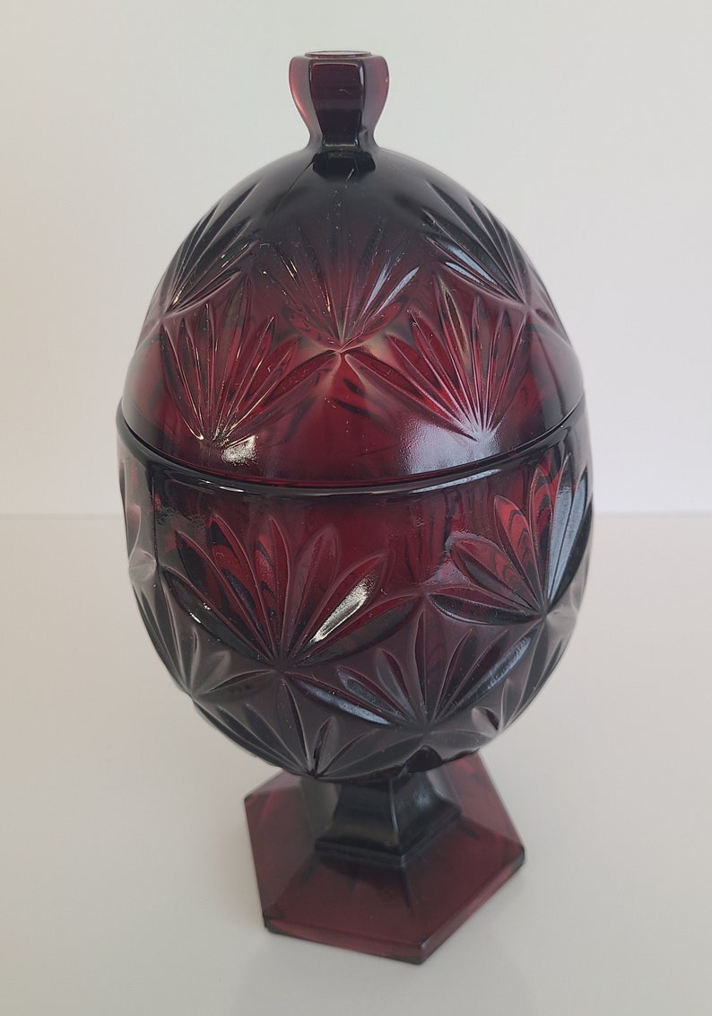 May include: A red glass candy dish with a lid. The dish is shaped like an egg and has a geometric pattern. The dish is on a pedestal.