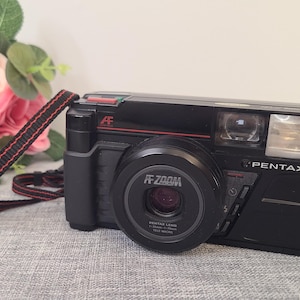 Op de afbeelding: Een zwarte Pentax AF Zoom camera met een rode band. De camera heeft een lens met de tekst "AF Zoom" en "Pentax Lens" erop gedrukt.