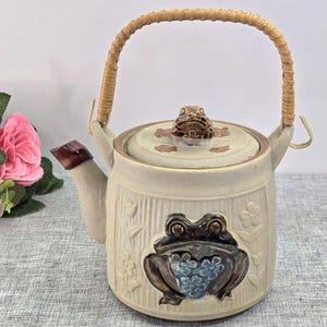 May include: A ceramic teapot with a light beige finish, featuring a prominent dark brown frog relief on the front and a smaller frog figurine on the lid. The teapot has a woven rattan handle and a spout with a dark brown tip, accented by floral patterns.