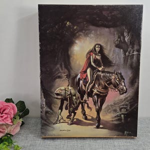 May include: Vintage fantasy art print featuring a woman in a red cape riding a black horse through a dark forest. A creature with a staff walks alongside. The artwork has a dark, gothic aesthetic, with detailed shading and a mysterious atmosphere.