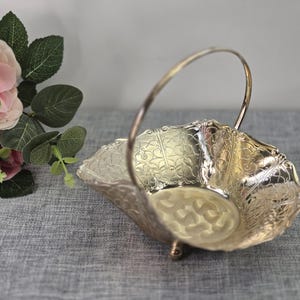 Può includere: Cestino decorativo argentato con manico. Il cestino presenta un motivo floreale in rilievo e un bordo smerlato. Il manico si inarca sopra il cestino. Il cestino è su una superficie grigia.