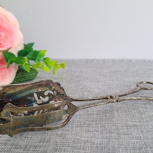 Puede incluir: Pinzas de servir plateadas con un diseño adornado y calado. Las pinzas están cerradas y sobre una superficie gris texturizada.