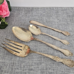 May include: Four silver-plated utensils, including a fork, two spoons, and a serving spoon, with ornate designs. The utensils are arranged on a grey textured surface.