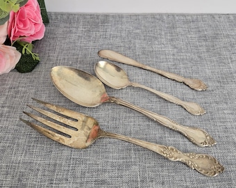 Könnte beinhalten: Vier versilberte Besteckteile, darunter eine Gabel, zwei Löffel und ein Servierlöffel, mit verzierten Designs. Das Besteck ist auf einer grauen, strukturierten Oberfläche angeordnet.